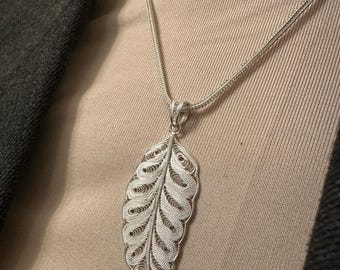 Op de afbeelding: Een zilveren filigraan blad hanger hangt aan een fijne ketting. De hanger is ingewikkeld ontworpen met gedetailleerde patronen, met een bladvorm. De ketting wordt gedragen over een neutraal gekleurde top en een donkergrijze blazer.