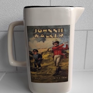 May include: White ceramic pitcher with a black rim and handle. The pitcher features a vintage Johnnie Walker label with illustrations of golfers and text that reads "Johnnie Walker" and "The whisky that goes with a swing."