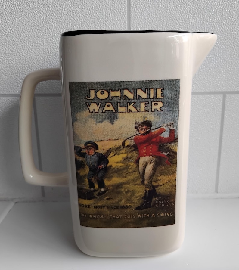 May include: White ceramic pitcher with a black rim and handle. The pitcher features a vintage Johnnie Walker label with illustrations of golfers and text that reads "Johnnie Walker" and "The whisky that goes with a swing."