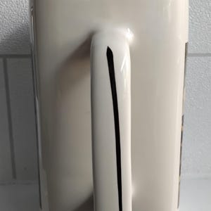 May include: A white ceramic mug with a rectangular shape and a curved handle. A black stripe runs down the center of the handle. The rim of the mug is black. The mug is on a white surface.