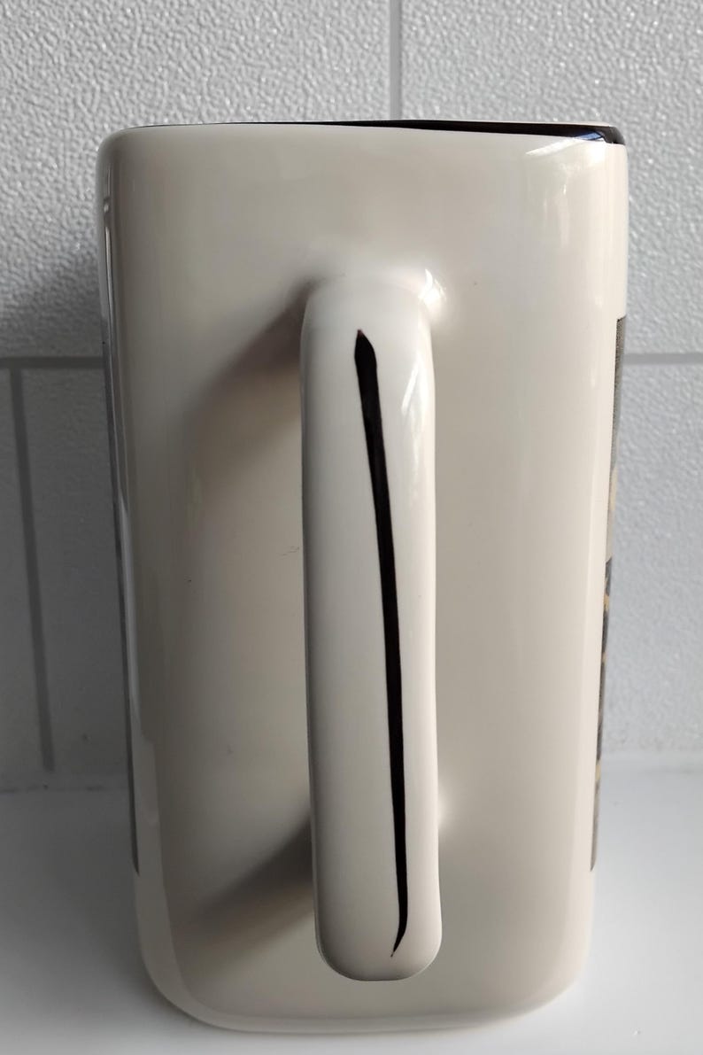 May include: A white ceramic mug with a rectangular shape and a curved handle. A black stripe runs down the center of the handle. The rim of the mug is black. The mug is on a white surface.