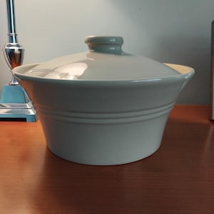 Puede incluir: Una fuente para horno de cerámica verde claro con tapa a juego. La fuente tiene forma redondeada con dos estrías horizontales en el centro. La tapa tiene un pequeño asa redondeada. La fuente está sobre una superficie de madera.