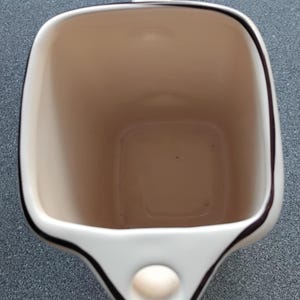 May include: A white ceramic mug with a square shape and a handle. The interior is beige, and the rim is outlined with a dark brown trim. A small, round, beige accent is on the front.