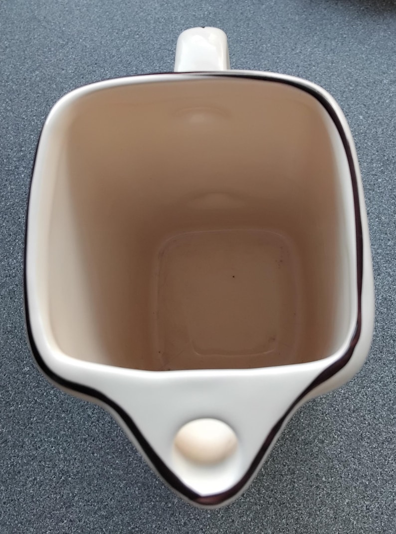 May include: A white ceramic mug with a square shape and a handle. The interior is beige, and the rim is outlined with a dark brown trim. A small, round, beige accent is on the front.