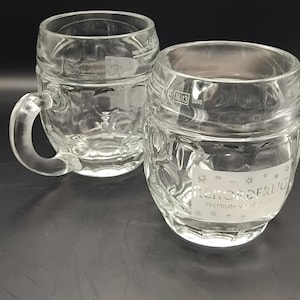 Könnte beinhalten: Zwei klare Bierkrüge aus Glas mit Griffen und strukturierten Seiten. Ein Krug steht aufrecht, der andere ist leicht geneigt. Die Krüge haben ein klassisches Design, das sich zum Servieren von Bier eignet. Der Text "Rekorderlig Premium Vart" ist sichtbar.