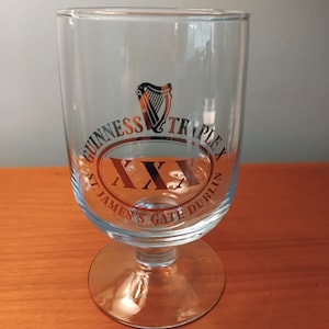Könnte beinhalten: Klares Guinness Triple X Bierglas mit rundem Fuß und geschwungenem Körper. Das Glas zeigt das Guinness-Logo mit einer Harfe und dem Text "Guinness Triple X St. James's Gate Dublin" in Orange und Schwarz.