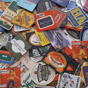 Puede incluir: Una gran colección de posavasos de cartón variados que presentan diversos logotipos de marcas, ilustraciones gráficas y textos, incluidos Underfall Lager, Fuller's Summer Ale, Carling Black Label, Murphy's Irish Stout y Coca-Cola, apilados densamente.