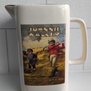 May include: Cream-colored ceramic pitcher with a handle and a spout. The front features a vintage advertisement for Johnnie Walker whisky, depicting two figures playing golf. Text includes "Johnnie Walker" and "The whisky that goes with a swing."