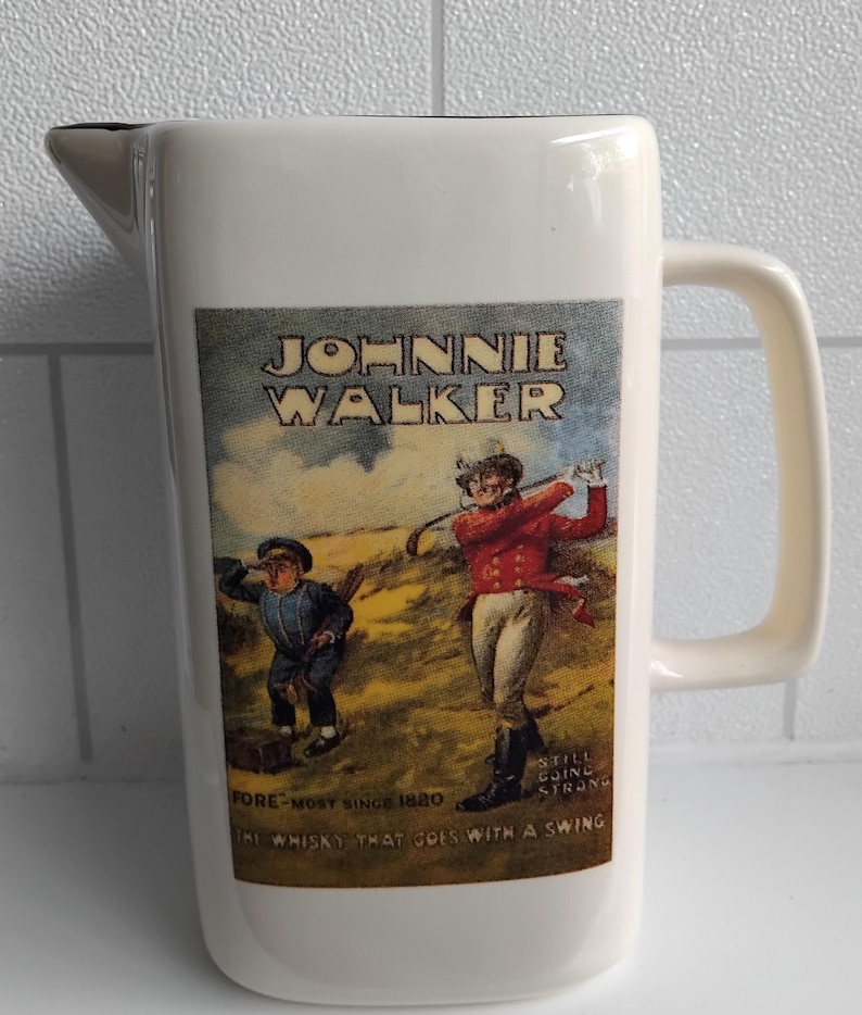 May include: Cream-colored ceramic pitcher with a handle and a spout. The front features a vintage advertisement for Johnnie Walker whisky, depicting two figures playing golf. Text includes "Johnnie Walker" and "The whisky that goes with a swing."