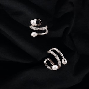 May include: Two silver ear cuffs with a pearl and crystal design. The cuffs are curved and have a delicate, feminine look.
