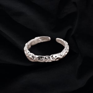 May include: A silver ring with a textured, organic design. The ring is open and adjustable.