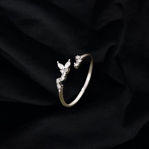 May include: A silver ring with a delicate design featuring a cluster of small, clear stones in the shape of leaves. The ring is open on one side, making it adjustable.