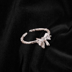 May include: A silver-toned adjustable ring with a bow-shaped design. The bow is adorned with clear, sparkling crystals. The ring has a textured band and is set against a dark background.