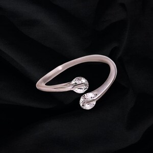 May include: A silver-toned cuff bracelet with two clear, round gemstones. The bracelet curves in a graceful, open design, with the gemstones set at the ends of the curved arms. The background is a dark, textured fabric.