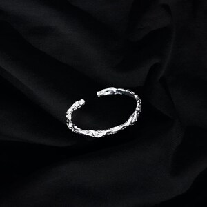 May include: A silver bracelet with a textured, organic design. The bracelet is open and has a simple, minimalist style.
