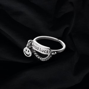 Pode incluir: Um anel de prata com uma corrente e um pingente que diz "Boa sorte" e tem um rosto sorridente.