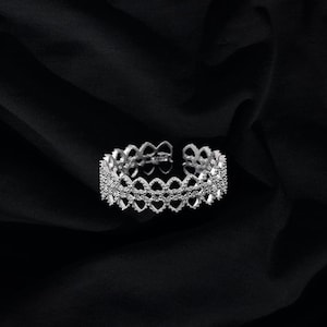 May include: A silver ring with a delicate, intricate design featuring multiple layers of small, connected circles. The ring is set against a black fabric background.