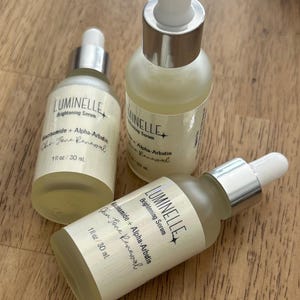 Puede incluir: Tres frascos cuentagotas de vidrio esmerilado de suero iluminador Luminelle, etiquetados como Niacinamide + Alpha-Arbutin Skin Tone Renewal, 30 ml. Los frascos tienen cuellos plateados y puntas de goma blancas, conteniendo un l&iacute;quido amarillo p&aacute;lido.