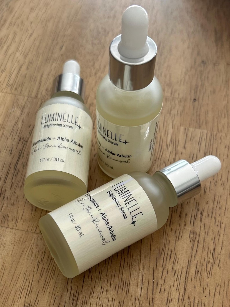 Puede incluir: Tres frascos cuentagotas de vidrio esmerilado de suero iluminador Luminelle, etiquetados como Niacinamide + Alpha-Arbutin Skin Tone Renewal, 30 ml. Los frascos tienen cuellos plateados y puntas de goma blancas, conteniendo un l&iacute;quido amarillo p&aacute;lido.