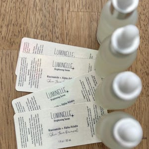 Puede incluir: Cuatro frascos cuentagotas de vidrio esmerilado dispuestos en una fila vertical, cada uno con una etiqueta blanca rectangular. Las etiquetas muestran el texto Luminelle Brightening Serum, Niacinamide + Alpha-Arbutin, Skin Tone Renewal y 30 ml.