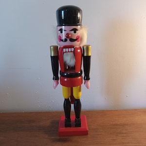 May include: A wooden nutcracker with a black hat, red and yellow clothing, and a white beard. The nutcracker has a painted face with pink cheeks, a black mustache, and a red nose.