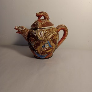 Op de afbeelding: Een rode en bruine theepot van keramiek met een drakenontwerp en ingewikkelde details. De theepot heeft een deksel met een drakenhandvat en is versierd met gouden en blauwe accenten.