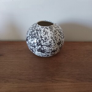 Puede incluir: Un jarrón de cerámica blanca con un diseño moteado en blanco y negro. El jarrón es redondo y tiene una abertura ancha.