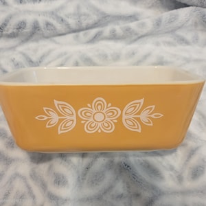 May include: A rectangular baking dish with a golden-yellow exterior and a white interior. The front side features a centered white graphic print of a stylized flower flanked by two symmetrical leaf patterns, all rendered in clean, simple lines.