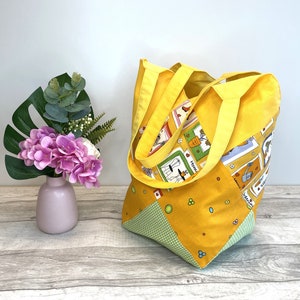 Könnte beinhalten: Eine gelbe Tragetasche mit einem grün-weiß karierten Muster am Boden. Die Tasche hat ein Patchwork-Design mit verschiedenen Mustern, die Bilder von Gartengeräten und Pflanzen zeigen. Die Tasche hat zwei gelbe Henkel.