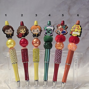 May include: Six decorative pens with character toppers. Each pen features a different design, including cartoon characters and house crests. The pens have colorful barrels and are displayed in a clear plastic holder.