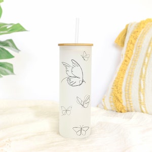 May include: A white frosted tumbler with a bamboo lid and a black line drawing of a butterfly. The tumbler has a straw and is decorated with several butterflies.