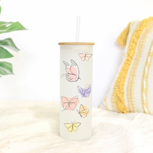 May include: A white tumbler with a bamboo lid and a straw. The tumbler has a design of butterflies in various shades of pink, purple, and yellow.