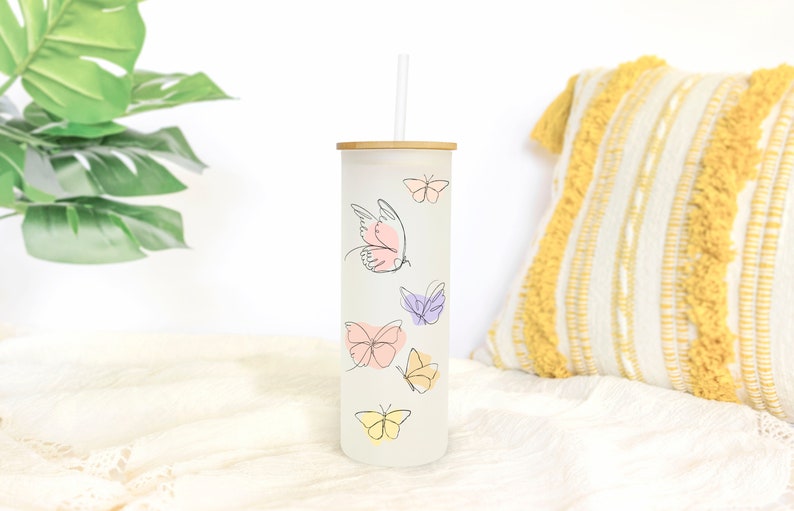May include: A white tumbler with a bamboo lid and a straw. The tumbler has a design of butterflies in various shades of pink, purple, and yellow.