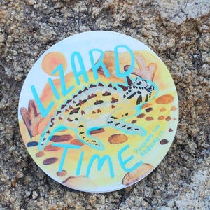 May include: A white round sticker with a yellow, orange, and brown lizard on a yellow, orange, and brown background. The text "Lizard Time" is written in blue on the sticker.