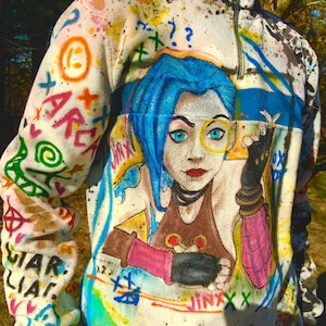 Può includere: Un pile bianco con un design a spruzzo di vernice colorata che presenta un personaggio dei cartoni animati con i capelli blu e il testo "Jinx" sul davanti.