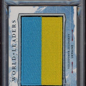 Puede incluir: Una tarjeta de colección Leaf Decision Vault 2022 con una foto de Volodymyr Zelenskyy, el presidente de Ucrania. La tarjeta tiene un parche azul y amarillo que representa la bandera ucraniana. La tarjeta está calificada por PSA y tiene una calificación de 9.