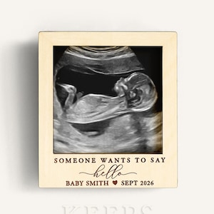 Puede incluir: Un marco de madera con una imagen de ecografía de un bebé. El marco tiene el texto "SOMEONE WANTS TO SAY hello BABY SMITH SEPT 2026". La ecografía es en blanco y negro.