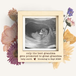 May include: A framed ultrasound image surrounded by dried flowers. The frame has text that reads "only the best grandma gets promoted to great grandma baby smith blooming in Sept 2026". The image is in black and white.
