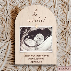 May include: A wooden pregnancy announcement frame with the text "hi auntie!" and an ultrasound image. The frame includes the text "I can't wait to meet you! Baby Anderson April 2024". A small wooden circle holds the ultrasound photo in place.