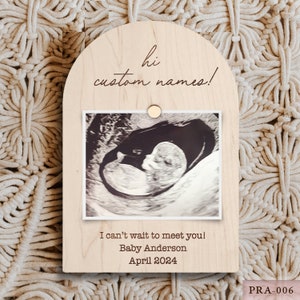 May include: A light-colored wooden plaque with an arched top, featuring a sonogram photo held by a small wooden circle. The plaque has the text "hi custom names!" and "I can't wait to meet you! Baby Anderson April 2024".