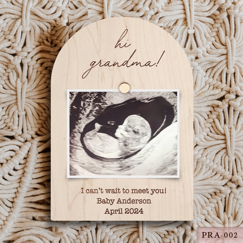 May include: A light-colored wooden plaque with an arched top, featuring the text "hi grandma!" and an ultrasound image. Below the image, it reads "I can't wait to meet you! Baby Anderson April 2024". A small wooden circle secures the photo.
