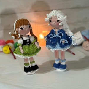 May include: Two handmade crochet dolls. One doll has brown hair in braids, a green dress, and black shoes. The other doll has white hair, a blue dress, and blue shoes. Both dolls have light skin and are standing on clear bases.