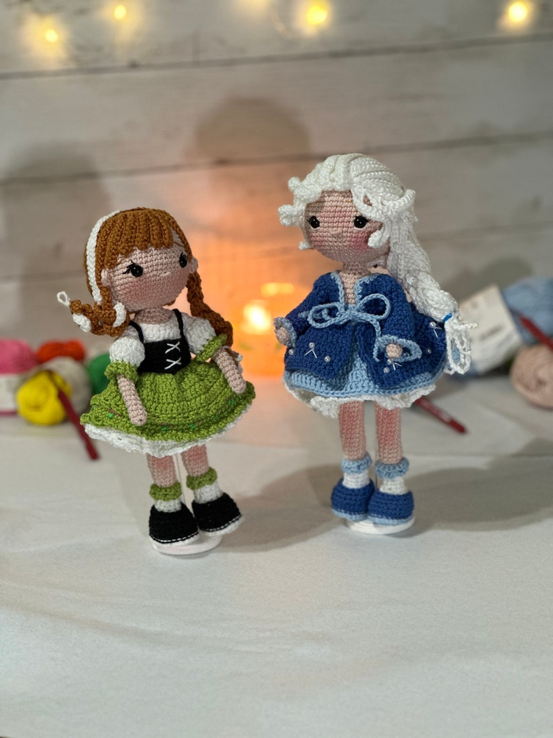May include: Two handmade crochet dolls. One doll has brown hair in braids, a green dress, and black shoes. The other doll has white hair, a blue dress, and blue shoes. Both dolls have light skin and are standing on clear bases.