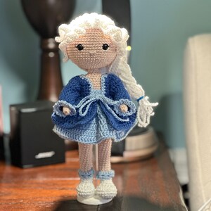 May include: A handmade crochet doll with a light skin tone, white hair, and a blue dress. The doll's dress is a mix of dark and light blue, with white accents. The doll is standing on a white base.