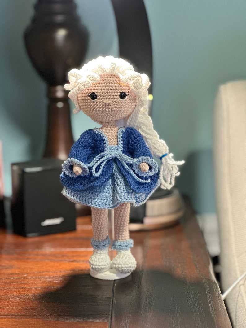 May include: A handmade crochet doll with a light skin tone, white hair, and a blue dress. The doll's dress is a mix of dark and light blue, with white accents. The doll is standing on a white base.