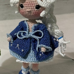 May include: A handmade crochet doll with a white wig and a blue outfit. The doll has a pink face with black eyes and is wearing a blue jacket with pearl accents. The doll's shoes are blue and white.