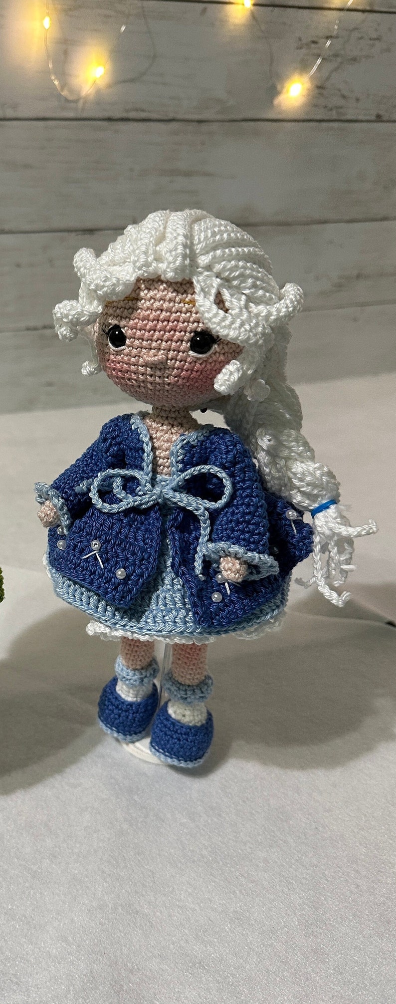 May include: A handmade crochet doll with a white wig and a blue outfit. The doll has a pink face with black eyes and is wearing a blue jacket with pearl accents. The doll's shoes are blue and white.