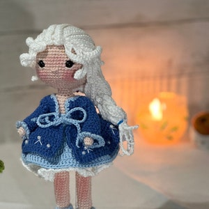 May include: A handmade crochet doll with a light skin tone, white hair, and a blue dress. The doll's dress is adorned with white accents and a bow. The doll is standing on a white surface.