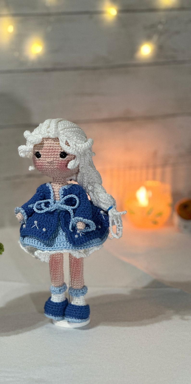 May include: A handmade crochet doll with a light skin tone, white hair, and a blue dress. The doll's dress is adorned with white accents and a bow. The doll is standing on a white surface.