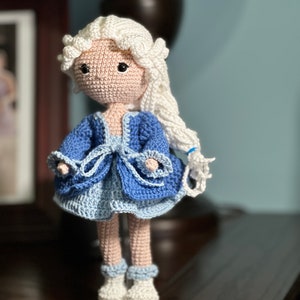 May include: A handmade crochet doll with a light skin tone, white hair, and black eyes. The doll wears a blue dress with a light blue skirt and white shoes. The doll is standing on a white base.